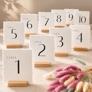 Peut inclure: Cartes de numéros de table blanches avec des chiffres noirs de 1 à 10, chacune sur un petit socle en bois. Un bouquet de fleurs séchées roses, beiges et vertes est au premier plan. Parfait pour les mariages ou événements.