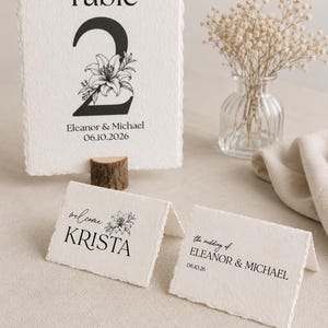 Peut inclure: Une carte de numéro de table rectangulaire aux bords effilochés, affichant le texte Table 2, une illustration florale, les noms Eleanor & Michael et la date 06.10.2026. Deux marque-places pliés sont inclus, dont un indiquant welcome KRISTA.