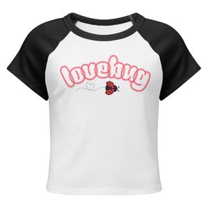 Pode incluir: Top curto branco com mangas pretas. A palavra "lovehug" em letras rosa bolha com contorno vermelho, uma joaninha e um coração pontilhado adornam a frente. Decote redondo.