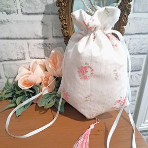 May include: A white drawstring bag with a pink floral pattern and a pink tassel. The bag has white ribbon straps and is sitting on a wooden surface. The background includes a mirror and faux flowers.