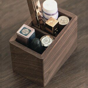 Può includere: Un organizer per trucchi in legno marrone scuro con più scomparti, pieno di vari prodotti cosmetici. L'organizer contiene articoli come rossetto, ciprie compatte e vasetti. L'estetica generale è pulita e ordinata.
