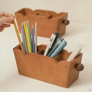 May include: Wooden desk organizer with a scalloped edge design. The organizer is filled with pencils, pens, and other writing utensils. A second organizer is visible in the background. The wood has a warm, natural finish.