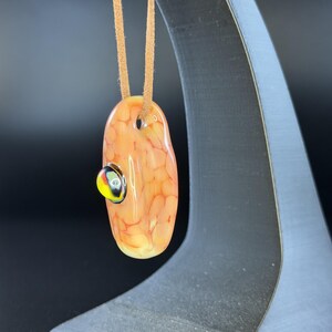 May include: An oval-shaped glass pendant featuring a mottled peach and orange pattern with a small, raised circular accent in black, yellow, and white. The pendant is suspended from a tan suede cord, creating a smooth, polished appearance.