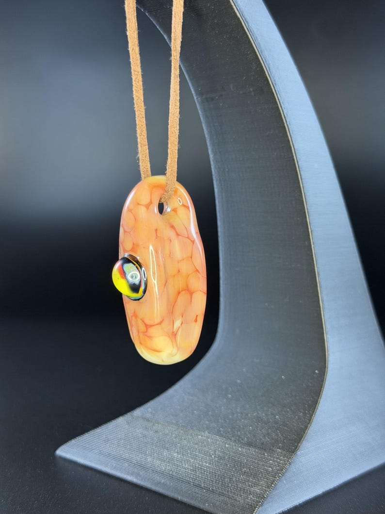 May include: An oval-shaped glass pendant featuring a mottled peach and orange pattern with a small, raised circular accent in black, yellow, and white. The pendant is suspended from a tan suede cord, creating a smooth, polished appearance.