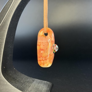 May include: A smooth, elongated glass pendant featuring an orange and cream honeycomb pattern. The pendant is suspended from a tan leather cord and includes a small, protruding glass accent piece with multicolored stripes on its side.