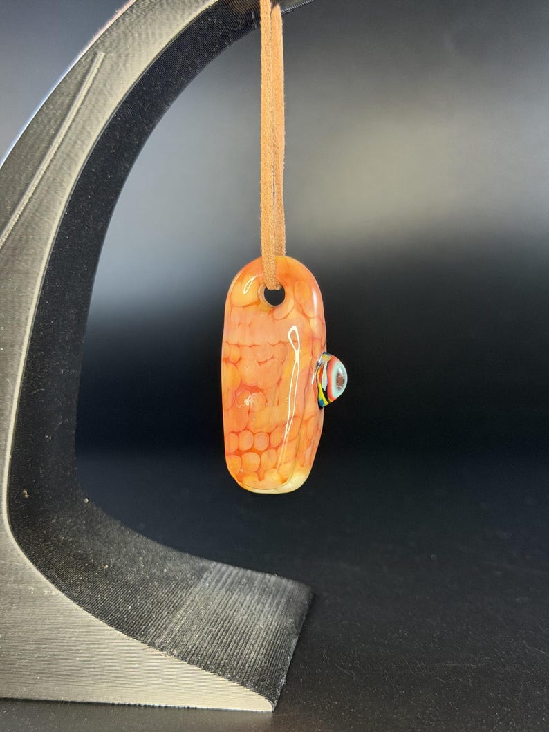 May include: A smooth, elongated glass pendant featuring an orange and cream honeycomb pattern. The pendant is suspended from a tan leather cord and includes a small, protruding glass accent piece with multicolored stripes on its side.