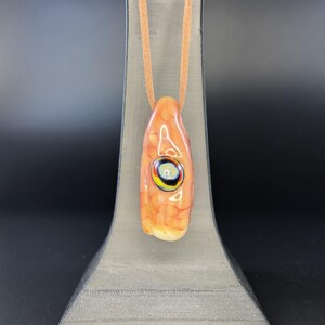May include: A glass pendant with a mottled orange and cream surface featuring a central eye-like design with concentric rings of black, yellow, and white. The pendant is suspended from a tan leather cord, creating a smooth, polished appearance.