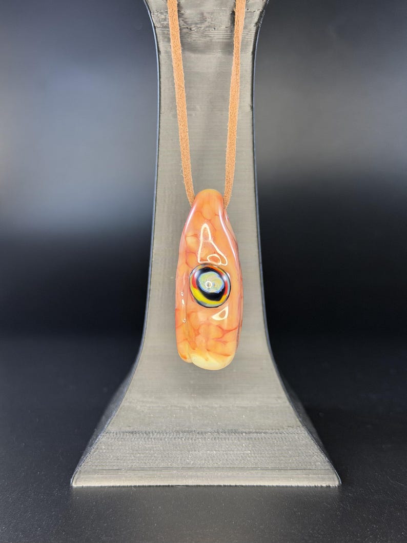 May include: A glass pendant with a mottled orange and cream surface featuring a central eye-like design with concentric rings of black, yellow, and white. The pendant is suspended from a tan leather cord, creating a smooth, polished appearance.
