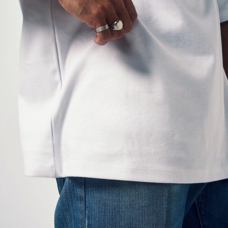 May include: Close-up of a white t-shirt with a crew neck. The shirt is paired with blue denim jeans. A hand with two silver rings is visible.