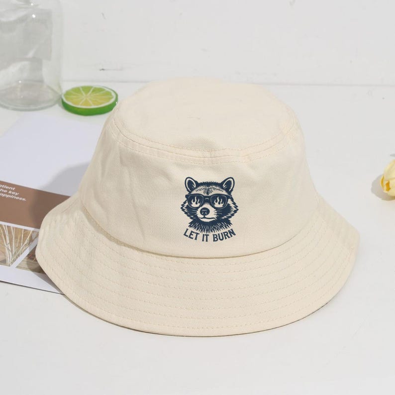 May include: Cream-colored bucket hat featuring a raccoon wearing sunglasses with the text "LET IT BURN" in navy blue. The hat has a wide brim and a rounded crown. A casual accessory for sun protection.