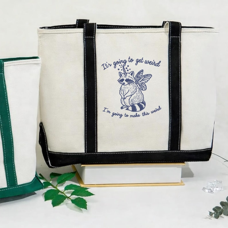 Puede incluir: Bolso tote color crema con ribetes y asas negras. El bolso presenta una ilustraci&oacute;n caprichosa de un mapache con un cuerno de unicornio y alas de hada, junto con el texto "It's going to get weird. I'm going to make this weird."