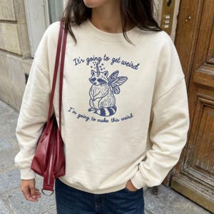 Puede incluir: Sudadera color crema con un gr&aacute;fico azul de un mapache con cuerno de unicornio y alas de hada. El texto "It's going to get weird" y "I'm going to make this weird" est&aacute;n impresos en azul.