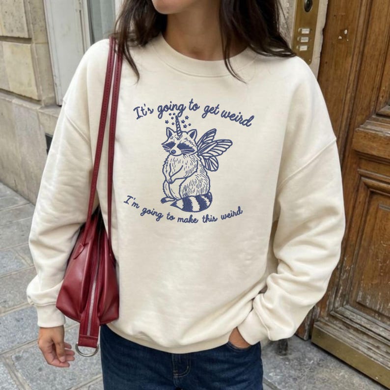 Puede incluir: Sudadera color crema con un gr&aacute;fico azul de un mapache con cuerno de unicornio y alas de hada. El texto "It's going to get weird" y "I'm going to make this weird" est&aacute;n impresos en azul.