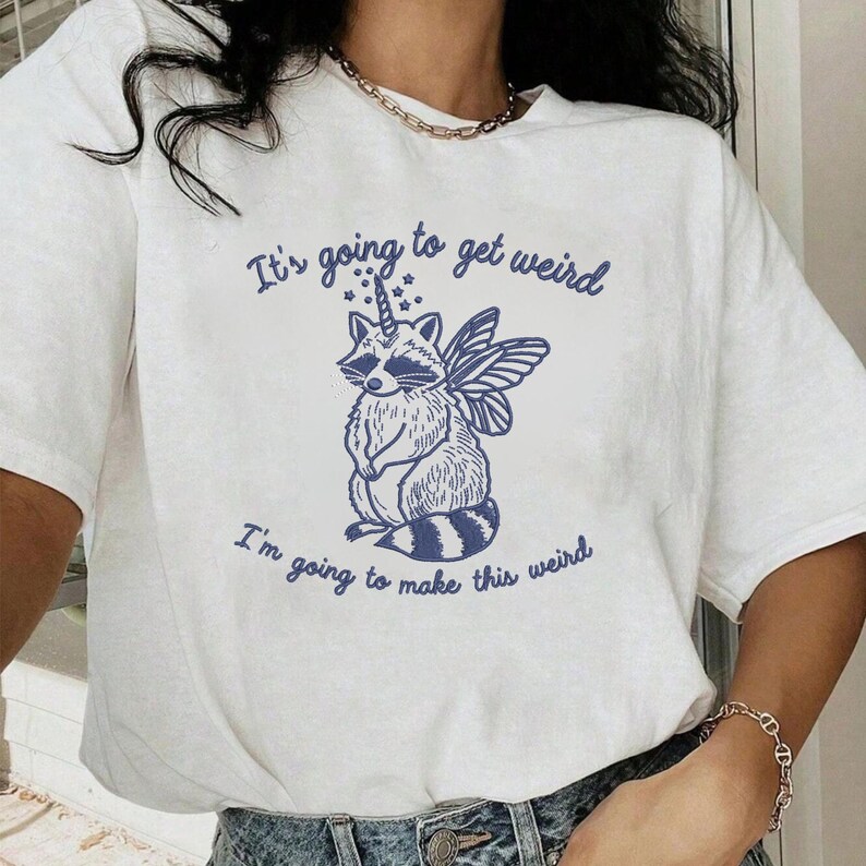 Puede incluir: Camiseta blanca con un dise&ntilde;o azul de un mapache con alas de hada y un cuerno de unicornio. El texto "It's going to get weird" y "I'm going to make this weird" est&aacute; impreso en azul encima y debajo de la ilustraci&oacute;n.