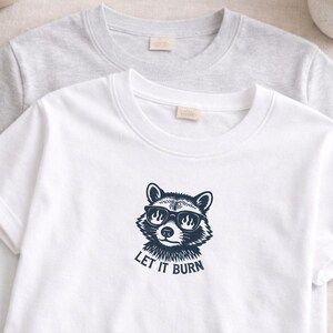 May include: Two crew neck t-shirts, one white and one gray. The white shirt features a navy blue graphic of a raccoon wearing sunglasses with flames in the lenses and the text "LET IT BURN" below.