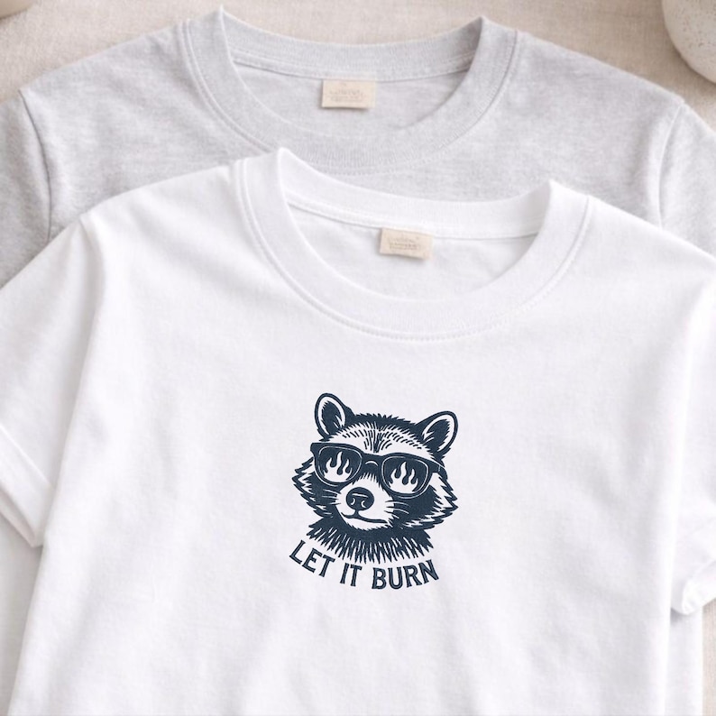 May include: Two crew neck t-shirts, one white and one gray. The white shirt features a navy blue graphic of a raccoon wearing sunglasses with flames in the lenses and the text "LET IT BURN" below.