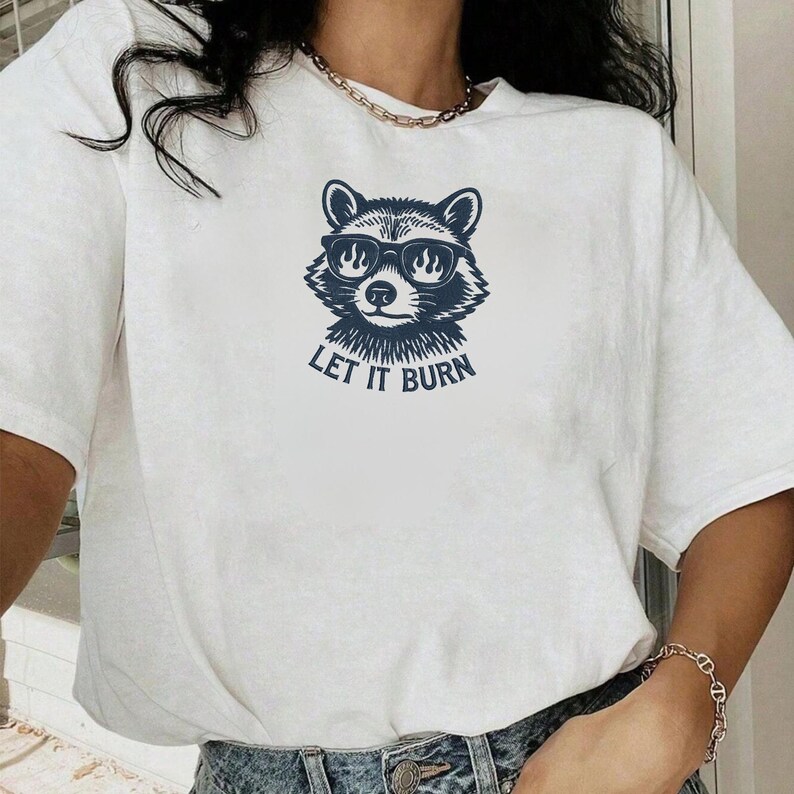 May include: White t-shirt featuring a navy blue raccoon wearing sunglasses with flames in the lenses. The text "LET IT BURN" is below the raccoon graphic. The shirt is a casual, unisex style.