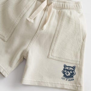 May include: Cream-colored shorts with an elastic waistband and drawstring. Features two front pockets and a raccoon graphic wearing sunglasses with the text "LET IT BURN" in navy blue. Casual wear.