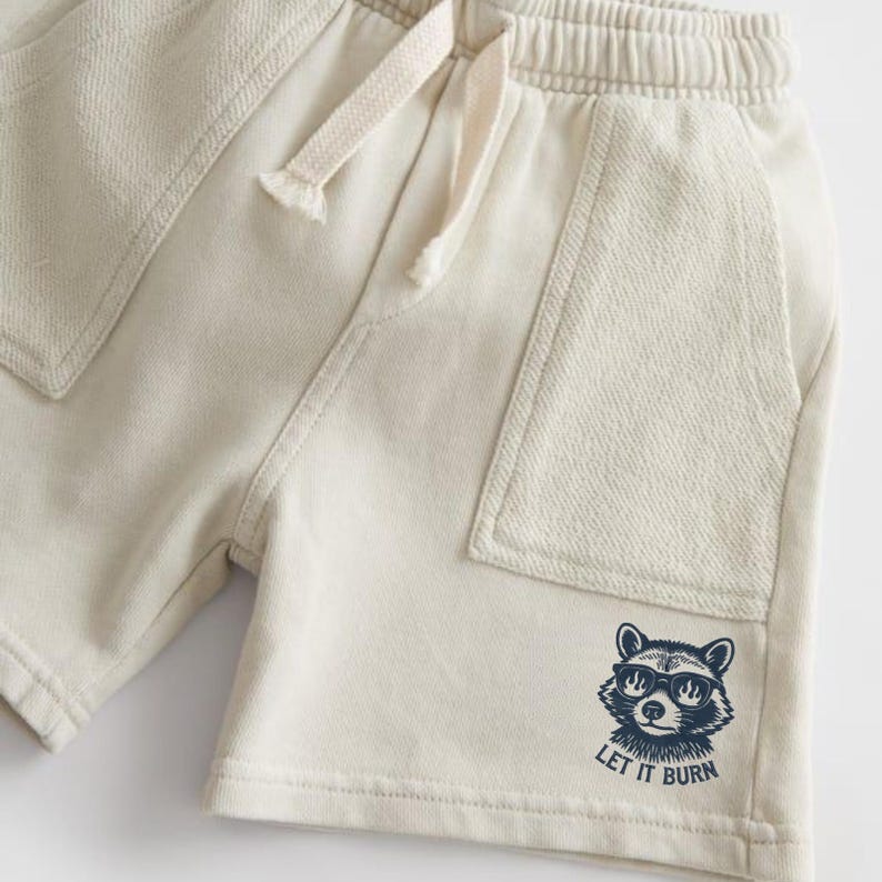 May include: Cream-colored shorts with an elastic waistband and drawstring. Features two front pockets and a raccoon graphic wearing sunglasses with the text "LET IT BURN" in navy blue. Casual wear.