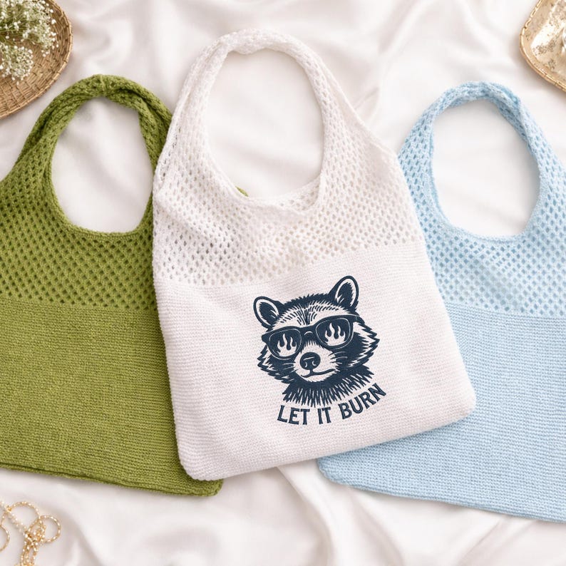 May include: Three knitted tote bags in green, white, and light blue. The white bag features a raccoon graphic with sunglasses and the text "LET IT BURN". The bags have a net-like texture and a single shoulder strap, ideal for carrying groceries or personal items.
