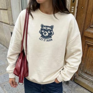 May include: Cream-colored sweatshirt featuring a raccoon graphic wearing sunglasses, with the text "LET IT BURN" in navy blue. The sweatshirt has a crew neck and long sleeves, suitable for casual wear.