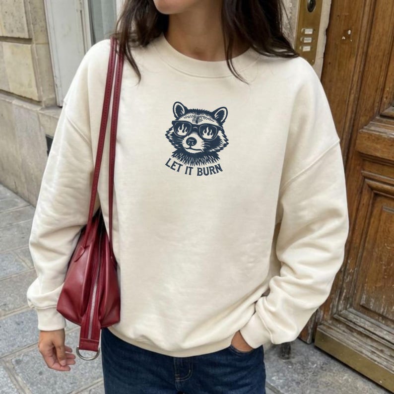 May include: Cream-colored sweatshirt featuring a raccoon graphic wearing sunglasses, with the text "LET IT BURN" in navy blue. The sweatshirt has a crew neck and long sleeves, suitable for casual wear.