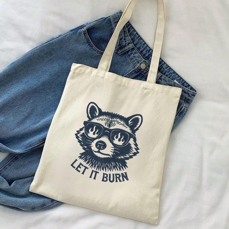 May include: Cream-colored tote bag featuring a raccoon wearing sunglasses with flames in the lenses. The text "LET IT BURN" is printed below the raccoon. The bag has two handles and is placed on top of a pair of blue jeans.