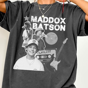 Puede incluir: Camiseta gris oscuro con el nombre "Maddox Batson" en letras blancas y una estrella. La camiseta incluye imágenes de un joven, un disco, una guitarra y una cinta de cassette. La palabra "PLATLIST" también es visible.