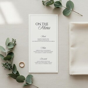 Może przedstawiać: Karta menu weselnego z napisem "ON THE Menu" w eleganckiej czcionce. Menu zawiera trzydaniowy posiłek: pieczoną zupę pomidorową, pierś z kurczaka i sernik waniliowy. Obecna jest również złota obrączka i gałązki zieleni.