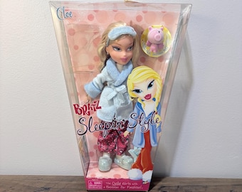 May include: A Bratz Sleeping Style doll in a clear plastic package. The doll has blonde hair, blue eye shadow, and is wearing a blue robe, eye mask, and pink pajamas. A small pink pig figure is included.