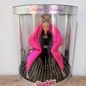 May include: A Barbie doll in a clear display case. The doll wears a black gown with silver accents and a pink shawl. The case has the words "Happy Holidays" and "Barbie" printed on it.
