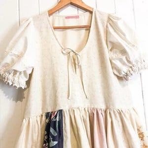 May include: A cream-coloured dress with short, puffed sleeves and a V-neckline. The dress features a patterned bodice and a tiered skirt with floral and patterned panels. It hangs on a wooden hanger against a white background.