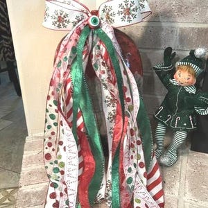 May include: A decorative Christmas bow featuring white ribbon with gold snowflake patterns, accented by long, cascading streamers in red, green, and white with polka dot, candy cane, and glitter textures, topped with a small green eye-like button.