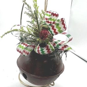 May include: A decorative Christmas bell with a red-brown finish, adorned with a green and red zigzag ribbon bow, faux pine needles, and a glittery red ornament. The bell is suspended by a gold-colored wire.