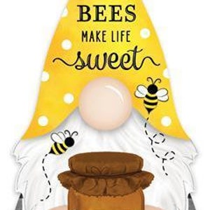 Op de afbeelding: Een grillige gnome-vormige wanddecoratie met een gele stippenhoed en een witte baard. De gnome houdt een pot honing vast en de tekst "BEES MAKE LIFE SWEET" is te zien. Het ontwerp bevat twee cartoonbijen.