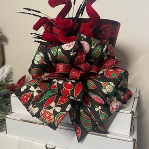 May include: A festive Christmas decoration featuring a black and red buffalo plaid container. The container is adorned with a large bow made of black fabric with red, green, and gold ornament designs. Red velvet accents and black sprigs add to the holiday theme.