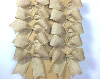 May include: Ten tan burlap bows arranged in two vertical columns on a rectangular cardboard backing. Each bow features a textured fabric finish with a central knot and two downward-facing tails, creating a uniform appearance across the set.