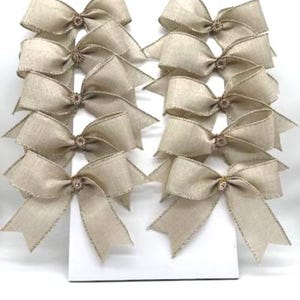 May include: A set of ten beige burlap bows arranged in two vertical columns. Each bow is crafted from a textured, light tan fabric with frayed edges and features a small, circular, metallic-toned embellishment at the center knot.