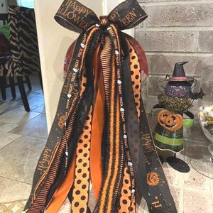 Może przedstawiać: Dekoracyjna kokarda na Halloween z wieloma wstążkami w kolorze czarnym i pomarańczowym. Wstążki mają wzory w czarne kropki na pomarańczowym tle, pomarańczowo-czarne paski oraz czarny materiał z napisem Happy Halloween i Trick or Treat.