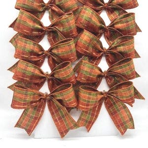 May include: A set of ten decorative bows arranged in two vertical columns. Each bow is crafted from a textured fabric featuring a plaid pattern in shades of burnt orange, forest green, and brown, with a visible weave and frayed edges.