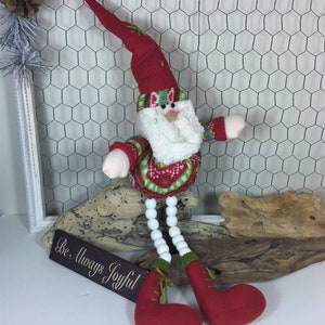 Op de afbeelding: Een rode en groene gebreide kerstmanpop met een witte baard en witte kralenbenen. De pop zit op een stuk drijfhout met een bord waarop staat "Wees altijd vrolijk".