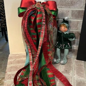 May include: A decorative Christmas bow featuring multiple layered ribbons in red, green, and plaid patterns, topped with a small button. Beside the bow stands a small elf figurine wearing a dark green velvet outfit with striped leggings.