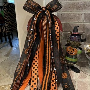Pode incluir: Um laço decorativo de Halloween com várias fitas penduradas em preto e laranja. As fitas exibem padrões que incluem bolinhas pretas em fundo laranja, listras laranjas e pretas e tecido preto com o texto Happy Halloween.