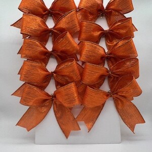 May include: A collection of orange decorative bows with a metallic sheen. The bows are arranged in rows, showcasing their symmetrical design and textured surface. These bows are suitable for gift wrapping or craft projects.