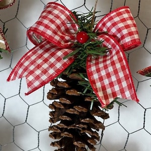 Puede incluir: Adorno navideño rústico con una piña grande, adornado con un lazo a cuadros rojo y blanco y vegetación artificial. Una pequeña baya roja añade un toque de color. El adorno cuelga sobre un fondo de malla metálica.