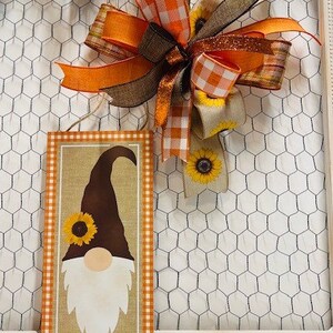 May include: A brown and white gnome with a sunflower on its hat is featured on a rectangular sign with an orange and white checkered border. The sign is hanging on a wire mesh background with a large orange and white bow.