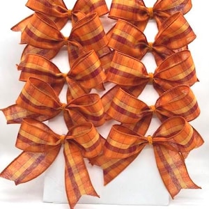 May include: A collection of ten decorative bows arranged in two vertical columns. Each bow is crafted from a translucent, textured fabric featuring a plaid pattern in shades of orange, yellow, and deep red, with two long trailing ribbon ends.