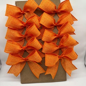 May include: Twelve orange fabric bows tied with a simple knot. The bows are arranged in four rows of three on a brown cardboard background.