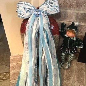 Könnte beinhalten: Eine dekorative Schleife aus weißem Band mit blauen Schneeflockenmustern, einem zentralen blauen Knopf und langen, herabhängenden Bändern in Hellblau, Weiß und Silber. Daneben steht eine kleine Elfenfigur in einem dunkelgrünen Outfit.