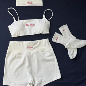 May include: A white matching set consisting of a cropped camisole with thin straps, high-waisted shorts, a headband, and a pair of socks. The camisole features the red embroidered text Mrs. Cecere, while the headband and shorts display the word Bride.